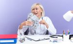 joyful-adult-slavic-female-doctor-medical-robe-with-stethoscope-sitting-desk-with-office-tools-holding-money-looking-side-isolated-purple-background